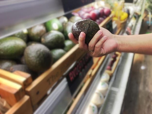 A person's hand selecting ripe avocados from a diverse selection of fruits and vegetables in a well-lit grocery store produce section, with other shoppers blurred in the background.